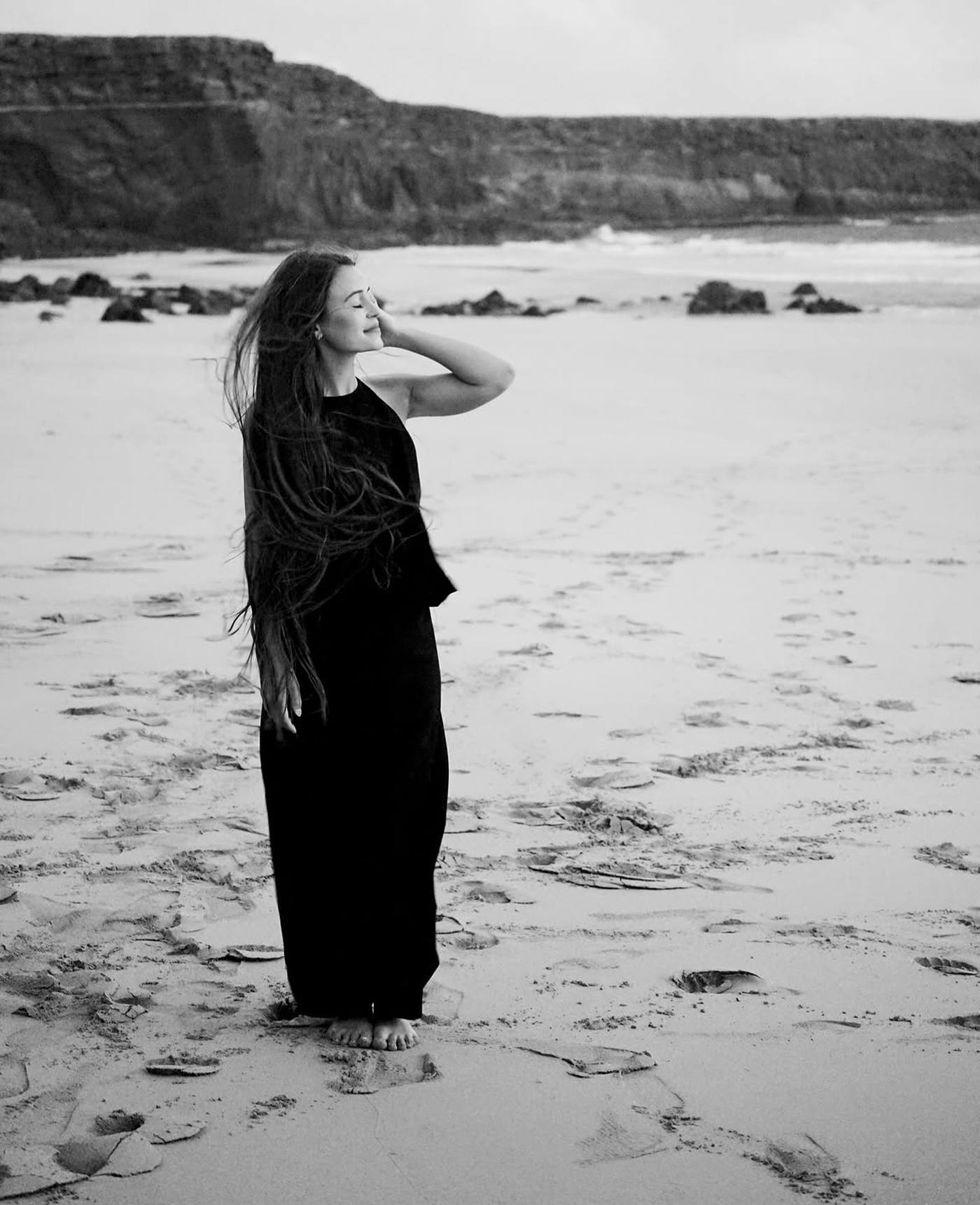 Portrait d'Elena Ruano, photographe, sur une plage, en robe noire, les cheveux longs au vent, les yeux fermés