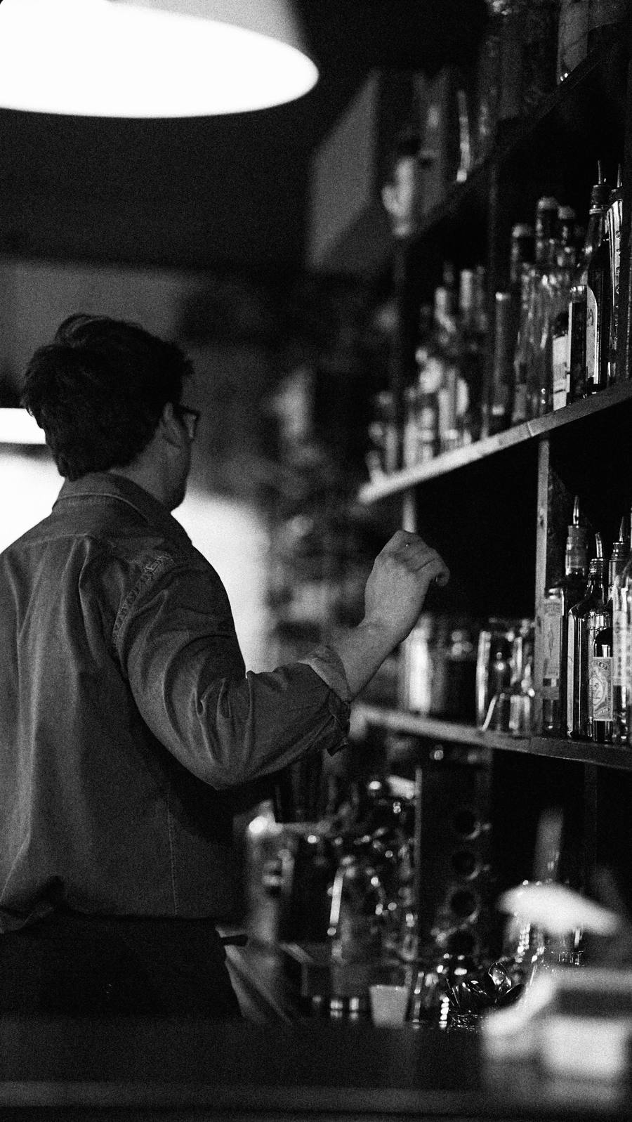 Un barman de dos, devant une étagère de bouteilles, sous une lampe suspendue