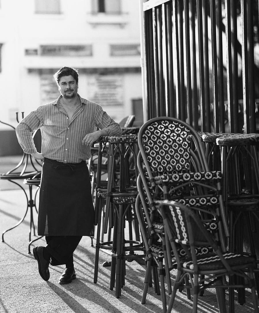 Un homme en chemise rayée et tablier, appuyé contre des tabourets de bistrot empilés au soleil