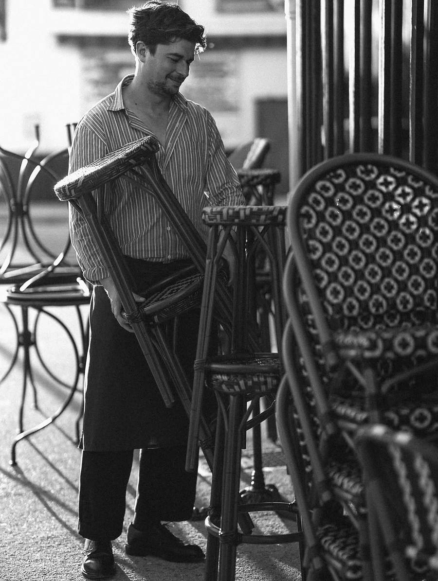 Un homme en tablier soulève un tabouret de bistrot et sourit, en lumière rasante